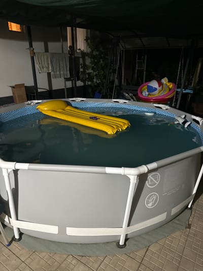 This media file shows an above-ground swimming pool set up outdoors at night. The pool has a grey exterior and a blue and white patterned interior liner. A yellow inflatable pool float is partially submerged in the water. To the left of the pool, a beach ball and an inflatable ring are suspended by chains, possibly as decorations or for play. In the background, there is some foliage and what appears to be a metal structure, perhaps a canopy or shelter. A cardboard box is visible on the ground near the pool. The scene is illuminated by artificial light, indicating it is nighttime. The ground surrounding the pool is paved with square tiles. The location is Pîrlița, Moldova.