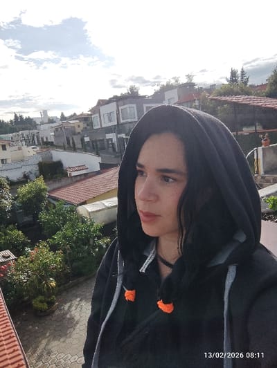 A young female subject is positioned in the foreground, facing right in a three-quarter profile, against a residential background in Puembo, Ecuador. She wears a black hooded jacket with a visible grey zipper lining and a dark choker necklace. Two small, spherical orange ornaments are attached to the jacket's drawstring. Her dark hair is partially visible beneath the hood. The background shows a multi-layered urban landscape featuring various residential buildings with predominantly white or light-colored facades and a mix of reddish-brown tiled roofs and flatter structures. Numerous green trees and vegetation are interspersed throughout the urban environment. A prominent white building with a steeple-like element is visible in the distant left. In the lower-left foreground, a red-tiled roof edge and a light-colored paved patio area are visible, along with dense green foliage. A decorative element, possibly an orange statue, is present on a wall in the mid-right background. The sky above is partly cloudy with significant white cloud cover. A digital timestamp of "13/02/2026 08:11" is embedded in the lower right corner of the image.