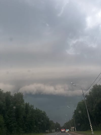 The image captures a view from a moving vehicle on a road, likely in the vicinity of Shelkovka, Russia, given the context. The sky is dominated by heavy, dark grey storm clouds, suggesting impending or ongoing inclement weather. A thick band of clouds stretches across the horizon, with lighter, more diffused clouds above.
Along the sides of the road, dense green trees line the route, creating a tunnel-like effect. The road itself appears to be asphalt and has at least one lane visible in the direction of travel. Streetlights with multiple bulbs are positioned along the right side of the road, some of which are illuminated. Electrical wires are strung between poles, also on the right.
A silver car with its brake lights illuminated is visible ahead in the same lane, indicating traffic. A white sign is present on the right side of the road, partially obscured by the trees. The overall impression is one of a somewhat ominous, overcast day, possibly during a summer storm, as one travels along a tree-lined highway. The image appears to have been taken with a mobile phone, with some minor smudges or water droplets visible on the lens in the upper portion of the frame.