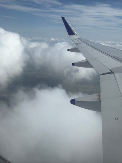An aerial view from an airplane window shows a wing of the aircraft extending into the frame from the right side of the image. The wing is white with blue accents on the wingtips. Below the wing, a landscape is visible through a layer of clouds. The clouds are dense and white, obscuring much of the ground. In some areas, the clouds are thin enough to reveal a patchwork of green fields and a winding river. A small cluster of buildings, likely a village or town, can be faintly seen nestled among the greenery. The sky above is a clear blue with wispy white clouds. The image appears to be taken during daylight hours. There are no people or specific landmarks clearly identifiable that would allow for definitive identification of the location as Mohan, India.