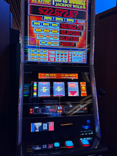 The image shows a close-up of two slot machines in what is likely a casino. The left slot machine displays "BLAZING JACKPOT WILD" and various jackpot amounts, including $22,672.67, $1,643.51, $1,073.13, and $250.38. The right slot machine shows the reel window with "5 J-A O-T" visible. A display shows $5,000 as the current cash amount. Control buttons such as "SPIN", "MAX BET," "BET ONE," and a pay table section are visible. The timestamp "09 42 05" is present. The image was taken in the city of Hollywood Seminole Reservation, United States of America.