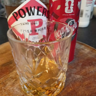 The image displays a close-up view of a glass containing amber liquid, likely whiskey, positioned on a wooden surface. Behind the glass, a bottle labeled "POWERS IRISH WHISKEY" is visible, featuring red and white branding. To the right of the whiskey bottle is a red Coca-Cola can with FIFA branding. The background is slightly blurred, showing hints of other items and a dark surface. The wooden surface appears to be a cutting board or table with visible grain and some scratches. The lighting suggests an indoor setting. The image does not contain any identifiable landmarks or contextual elements that would suggest a specific location within the city of Limerick, Ireland, or any other city. The scene depicts a still life arrangement of beverages.
