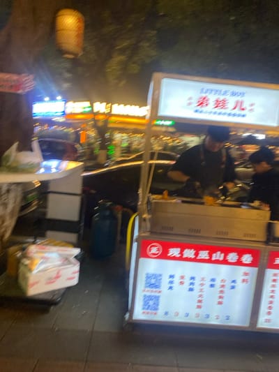 This image depicts a street food stall operating at night in 天宫殿街道, China. The scene is illuminated by artificial lights emanating from the food cart and surrounding commercial establishments.
In the foreground, a portable food cart with an illuminated canopy is visible. The top sign on the canopy reads "LITTLE BOY" alongside Chinese characters "弟娃儿". Below, a red banner displays "现做巫山卷卷" (Freshly Made Wushan Rolls), and further down, illuminated panels present two QR codes for payment or information, and a menu listing various food items or ingredients such as "加料" (add-ons), "鸡翅" (chicken wings), and "香辣鸡腿" (spicy chicken drumstick).
Two individuals, presumably vendors, are positioned behind the food cart. One individual, wearing a dark shirt and a dark cap, is leaning over a cooking surface, actively preparing food. The second individual is partially visible to the right. A blue gas cylinder connected by a yellow hose is situated on the ground behind the cart. To the left of the food cart, a white stand holds a white plastic bag and several stacked white boxes or packages, possibly takeout containers.
The background reveals a blurred street scene with vehicle lights and brightly lit commercial buildings featuring prominent orange and blue signage. A traditional-style, cylindrical lantern hangs from an overhead tree branch on the far left. The ground is a paved surface, likely a sidewalk.
