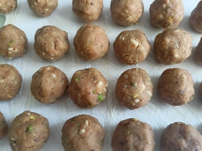 The image shows a close-up, overhead view of numerous round, light brown food items, likely dough balls or candies, arranged in rows on a white surface, possibly a table covered with plastic wrap. The food items appear to be made of a granular mixture, with visible flecks of lighter and darker ingredients, such as nuts and spices. They are not perfectly spherical, suggesting they have been hand-formed. Some of the food items have a slightly uneven texture and surface. The lighting is bright and diffused, casting minimal shadows, indicating an indoor setting with overhead light. There is no visible text in the image, nor are there any people or obvious location cues beyond the likely indoor setting. The overall impression is one of preparation or presentation of food.