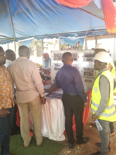 A group of six men stand around a table covered with a white tablecloth. One man, wearing a white shirt and a red vest with a red cross printed on it, stands to the left of a white board displaying photos, seemingly pointing at one. Three men stand to the right of the board, one wearing a bright yellow vest, the other in a blue shirt, and the last in a white hard hat with a white vest. Another man with a red vest is partially visible behind the board. The men are gathered under a tent, and a red tablecloth is visible to the right. The tent is open, and a small patch of green grass is visible behind the table. There is an outdoor setting behind the tent. The scene likely depicts a meeting or event related to the International Committee of the Red Cross (ICRC), as evidenced by the red cross logo worn by some participants. The location is Bunia, Democratic Republic of the Congo, specifically at coordinates latitude: 1.556, longitude: 30.244.