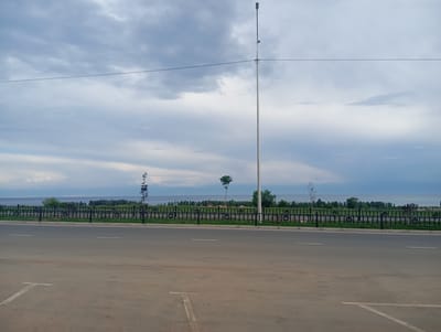 The image depicts a serene outdoor scene. In the foreground, a paved road with painted white lines suggesting parking spaces is visible. A black metal fence runs parallel to the road, separating it from a grassy area. Beyond the fence, a body of water, likely a lake, stretches across the horizon. The far shore is lined with a dense array of trees.
The sky is predominantly overcast with dramatic, textured clouds in shades of grey and white, suggesting either early morning or late afternoon light. A tall, thin lamppost stands prominently on the right side of the frame, its light fixture positioned at the very top. A thin wire stretches horizontally across the sky, connecting to the lamppost.
There are several trees visible on the grassy area between the fence and the water, including a distinctive, small tree with a rounded canopy located near the center. The overall atmosphere is peaceful and tranquil. The weather appears to be mild, with no obvious signs of rain or strong winds. The location cues, such as the trees and the body of water, are consistent with a lakeside setting in Kyrgyzstan. No people are visible in the image.