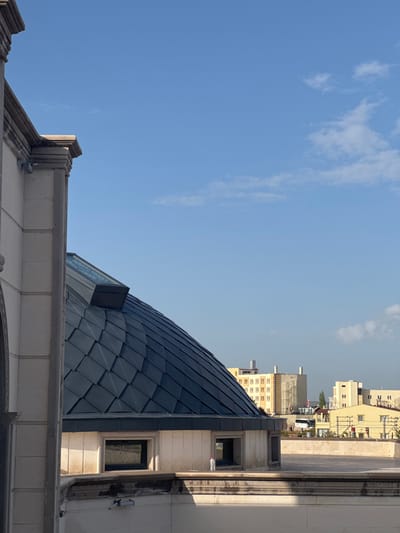 The image shows a low-angle view of building architecture in Ürgüp, Turkey, dominated by a blue-tiled dome and surrounding structures. The left side of the frame is filled with a clear blue sky with scattered clouds. The dome is constructed of individual, grey-blue tiles, forming a scaled pattern. Adjacent to the dome is a building with white stone or stucco facade, small windows, and architectural details. In the background, additional buildings are visible.