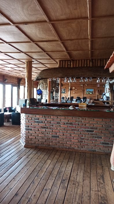 A rustic interior features a bar with a brick front and dark wood countertop. Wine glasses hang above the bar. The back of the bar shows wooden walls, decorative items, and people. One side features a smooth wood panel wall with support beams. The opposite side has a dark wood plank wall and floor. Sunlight streams in from a window below. The overall aesthetic is warm and inviting.