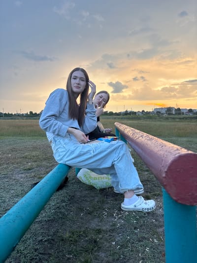 The image shows two young women sitting on a painted metal bar, likely a piece of playground equipment, outdoors at dusk. The woman in the foreground, with long brown hair and wearing a grey sweatshirt and ripped light-wash jeans, is looking towards the camera and touching her hair. She is wearing white Crocs. The woman behind her, wearing a black top, is partially visible and making a peace sign with her fingers. They are sitting on a blue and red painted bar with a grassy field and some distant buildings in the background under a cloudy sky with the sun setting, casting an orange glow. The location is specified as Sarotski Selski Sovet, Belarus.