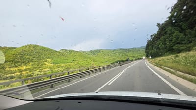 The image shows a view from inside a vehicle driving on a highway. The road has two lanes in each direction, separated by a double white line. The highway is flanked by a guardrail on the left and a steep, tree-covered hillside on the right. The landscape is characterized by rolling green hills covered in dense vegetation. The sky is overcast with some dark clouds, suggesting it might be raining or has recently rained, as evidenced by the water droplets on the windshield. In the distance, a truck and a signpost are visible on the right side of the road. The location is identified as the highway near Boița, Romania.