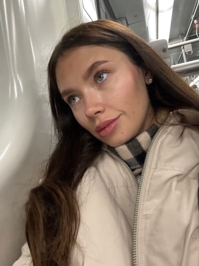 A close-up image portrays a female individual from the chest up, positioned within an indoor environment. She possesses long, dark brown hair, blue eyes, and light skin with several small moles on her right cheek. A small, circular, silver-colored stud earring is present in her right earlobe. Her lips are slightly parted, exhibiting a pinkish hue. She wears a light beige or off-white zip-up jacket, partially revealing a plaid scarf underneath with a pattern of black, white, and tan stripes. Her gaze is directed towards the upper left, away from the camera. The background on the left consists of a light-colored, possibly white, paneled or curved surface with vertical light reflections. To the upper right, metallic fixtures, potentially handrails or overhead structures, are visible alongside indistinct overhead lighting. The specific location within the Municipal Unit of Elliniko, Greece, cannot be ascertained from the visual evidence in the image.