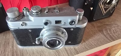 This is a close-up, eye-level shot of a vintage Zorki C film camera resting on a wooden surface. The camera, with its silver and black body, is the primary subject. The Zorki C logo is visible in Cyrillic script on the top of the camera. The lens, with various settings and the serial number "N8038419" and "F=5cm 1:2", is also clearly visible. Several dials and buttons adorn the top of the camera, including one with markings up to "30". To the right of the Zorki, a portion of another vintage camera, possibly a "BOX" camera, is visible. The background is out of focus, showing a blurred red fabric on the left and a wooden surface with a light grain. The lighting appears to be indoor, with no specific cues for time of day or weather. The scene suggests a collector's display or a personal collection of vintage photographic equipment. The overall impression is one of nostalgia and appreciation for classic technology.