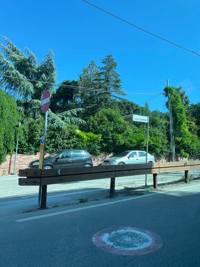 The image depicts an outdoor street scene under a clear blue sky on a bright, sunny day. The location is identified as Via Carmelo Colamonico in Cassano delle Murge, Italy, indicated by a white rectangular street sign with blue text positioned mid-ground on the right.

In the foreground, a dark wooden barrier, constructed from two horizontal planks affixed to vertical metal posts, extends across the lower portion of the image. The road surface is asphalt, featuring faded white dashed lines and a circular road marking with a red outer ring and a white inner area, which is worn.

To the left of the wooden barrier, a tall silver pole supports a circular "no entry" traffic sign (red with a horizontal white bar) and two smaller signs below it, one yellow and one blue. Behind the barrier and signs, a low, reddish-brown brick wall is visible.

Two automobiles are parked on the left side of the road, behind the barrier and against the brick wall. The vehicle closer to the foreground is a dark-colored (appears black or dark grey) hatchback. Further down, a silver-colored hatchback is parked. Both vehicles appear unoccupied.

The background is dominated by dense green foliage, including tall evergreen trees with prominent needle-like leaves and various bushes. A utility pole, partially overgrown with ivy or climbing plants, stands on the far right side of the image, with overhead wires extending across the blue sky. Bright sunlight casts distinct shadows on the road and under the barrier. No individuals are visible in the scene.