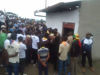 A crowd gathers near a small, simple building with a red door. Many individuals wear scout uniforms, some with neckerchiefs. Adults and children are present. The setting appears to be outdoors, possibly a rural area. A vehicle is visible in the background. The sky is overcast. The overall atmosphere suggests a community event or gathering.