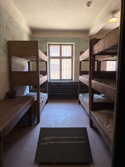 The image depicts the interior of a room at the Auschwitz-Birkenau Memorial and Museum in Oświęcim, Poland. The room contains two sets of wooden bunk beds, each with two levels, arranged along the walls. The beds are sparsely furnished with thin mattresses covered in a light-colored fabric, presumably to represent the conditions of the time.
On the left side of the room, a long wooden table or bench is positioned in front of a bunk bed. A woven basket sits on the table. The walls of the room have a faded, textured finish, suggesting the passage of time and wear. A large, multi-paned window with metal bars is visible in the background, offering a glimpse of the exterior. The floor is a smooth, muted color with some visible cracks.
In the foreground, a dark, angled informational plaque is placed on the floor. The plaque bears text in both Polish and English, indicating that it provides information about the room's furnishings and its use by inmates in Auschwitz I from 1941. The overall impression is one of starkness and austerity, reflecting the historical context of the location. A light fixture is visible hanging from the ceiling, and a smoke detector is mounted near the ceiling. The scene is lit by natural light from the window and possibly artificial light from the fixture.