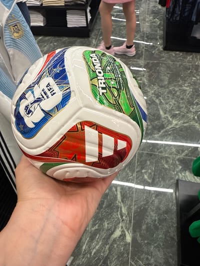A light-skinned hand holds a new, multicolored soccer ball indoors within a retail establishment in Sunrise, United States of America. The soccer ball, adorned with the FIFA logo and the word "TRIONDA," alongside text indicating "REPLICA MATCH BALL" in multiple languages, features an Adidas logo (three stripes) on one of its red and gold panels. Its design incorporates white, blue, green, and red/gold panels, visually consistent with the Adidas Al Hilm ball from the 2022 FIFA World Cup. The ball's surface appears glossy, potentially from a protective film. In the immediate background to the left, a light blue and white striped fabric with a gold crest, resembling an Argentina national football team jersey, is partially visible. Further back, black display shelves hold neatly folded light-colored merchandise. To the right, the lower legs of a person wearing pink athletic shoes, dark socks, and the bottom edge of a pink garment (skirt or shorts) are visible standing on a dark, glossy marble-patterned floor with white veining and reflections from overhead lighting. A black retail display unit and a partial green object are also present on the right side of the frame.
