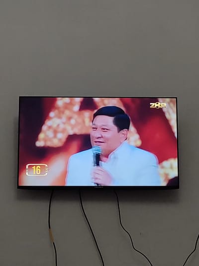 A television screen mounted on a wall displays a man holding a microphone. The man is wearing a white collared shirt. A graphic in the lower left corner of the screen shows the number "16" within a yellow rectangular box. In the upper right corner of the screen, a logo with the letters "ZOR" and a soccer ball is visible. The television is connected to black cables that hang down the wall. The wall appears to be painted a light gray color. The location is identified as Gurlan, Uzbekistan.