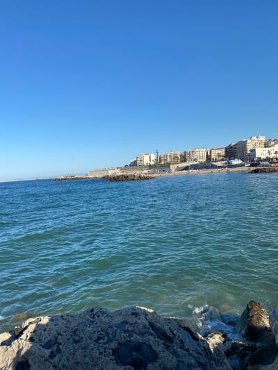 The scene is a coastline in Beau Fraisier, Algeria. The image depicts the shoreline with the city's buildings lined up along the coast. The buildings are medium to high-rise, light-colored structures. A road runs parallel to the shoreline, appearing to have some traffic and pedestrians. The sea is a light blue-green with gentle waves. The foreground shows a rocky embankment where the water meets the land. The sky is a clear, bright blue.