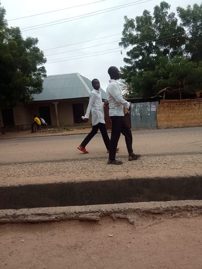 In the city of Biliri, Nigeria, two young men dressed in white long-sleeved shirts and dark trousers are walking along a paved road. The man in the foreground is wearing sandals, while the man slightly behind him is wearing red slippers. Both appear to be carrying items in their hands. To the left, a building with a corrugated metal roof is visible, with two figures seated on a bench in front of it. Across the road, there's a structure with a thatched wall. Overhead, power lines stretch across the sky. The road is separated by a concrete gutter or drain. The overall environment appears to be a residential or mixed-use area. The sky is overcast.