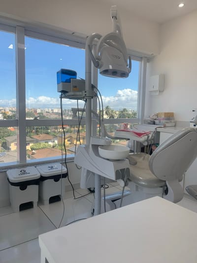 The image displays a clean, well-lit dental operatory in Lauro de Freitas, Brazil, during daylight hours. The room features white walls and a light-colored tiled floor. A modern, light grey and white dental chair is positioned centrally on the right, accompanied by its integrated unit. This unit includes a multi-LED dental operating light suspended above the chair, a white ceramic spittoon, and a blue-tinted water reservoir mounted on a vertical pole. Various hoses and wires extend from the equipment. To the right of the dental chair, a white countertop holds assorted dental supplies in clear organizers, with a white paper towel dispenser mounted on the wall above. A white table partially occupies the bottom right foreground. Against the window on the left, two white, foot-pedal operated waste bins are present: one labeled "LIXO COMUM" (Common Waste) with a recycling symbol, and the other "INFECTANTE" (Infectious) with a biohazard symbol. The large window offers a panoramic view of Lauro de Freitas under a clear blue sky with scattered clouds. The cityscape primarily consists of residential buildings with terracotta-tiled roofs, some featuring solar panels, interspersed with green vegetation and distant urban structures. No individuals are visible within the scene.
