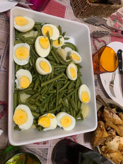 The image depicts a close-up view of a festive meal laid out on a table. The central focus is a white rectangular dish overflowing with cooked green beans and interspersed with sliced hard-boiled eggs. Several whole potatoes are also visible beneath the beans. Scattered throughout the dish are individual pieces of cooked chicken.

To the left of the green bean dish, a woven basket is partially visible, containing what appears to be food items. Next to the basket, a pair of silver tongs rests on the tablecloth. Further left, a glimpse of a reddish-purple glass object can be seen.

To the right of the green bean dish, a dark glass bottle, possibly wine, stands upright. Adjacent to it, a portion of a green glass is visible, reflecting light. In the background, a plate with a sliced roasted chicken is present, accompanied by a fork and knife. Another plate with cutlery and an orange glass filled with a beverage are also visible.

The table is covered with a patterned tablecloth featuring floral motifs. The overall impression is that of a shared meal, likely a dinner or lunch, given the abundance of food and the presence of drinks and serving utensils. The lighting suggests an indoor setting, possibly during the daytime or early evening. No discernible text is visible in the image. The scene evokes a sense of warmth and communal gathering.
