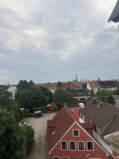 This is a high-angle, wide shot of a residential area in Bremerhaven, Germany, under a cloudy sky. The scene depicts a collection of multi-story buildings with red tile roofs, interspersed with mature trees and greenery. In the foreground, a bright red building with white-trimmed windows is prominent. To the left, a paved area contains a parked white van and a dark trailer. Further into the background, more buildings of varying architectural styles are visible, along with a church spire and distant city structures. The overall impression is of a settled urban neighborhood.