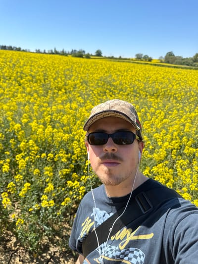 The image is a bright, sunlit selfie featuring a young man against a vibrant backdrop of yellow flowers. The perspective is taken from slightly above, with the man's head and torso occupying the right side of the frame. He is looking directly at the camera, wearing dark sunglasses that obscure his eyes. On his head is a beige baseball cap, patterned with what appears to be a Gucci GG monogram and signature red and green stripes. White wired earphones are visible in his ears, with the wire extending across his chest. He wears a dark t-shirt, possibly black or dark grey, featuring a graphic design with yellow text and a checkered flag motif, suggesting a racing or automotive theme. A dark strap from a bag is also visible across his shoulder.
The background is dominated by a vast field of bright yellow flowers, characteristic of rapeseed or canola, stretching into the distance. A line of trees marks the horizon where the field meets a clear, bright blue sky. The strong sunlight indicates it is daytime, likely midday or early afternoon, under sunny weather conditions. The immediate foreground on the right shows a patch of disturbed earth or a dirt path. The overall setting suggests a rural or semi-rural environment, consistent with the provided location context of North Hertfordshire, United Kingdom, during a time when such fields would be in bloom. The man appears relaxed, enjoying the outdoor scenery.