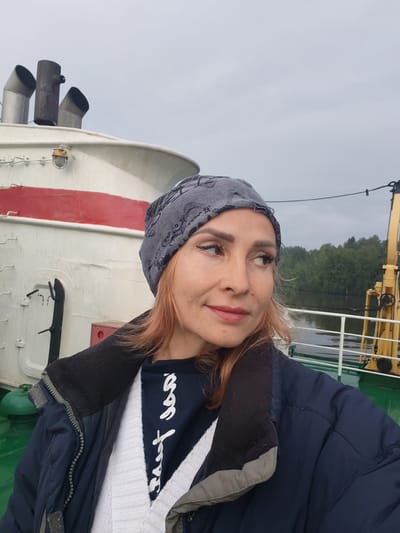 In an outdoor, overcast scene on a ship, a woman with reddish-blonde hair, wearing a grey patterned beanie, a dark jacket over a white knitted garment and a dark blue shirt with white lettering, is pictured from the chest up, looking to her right. The background features the white and red superstructure of a ship, with three metal funnels visible at the top. A white railing and yellow equipment are also present on the deck. The location is identified as the city of Shindalovo, Russia.