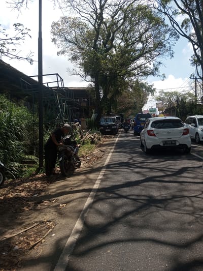 The image shows a street scene in what appears to be a tropical location. A man dressed in dark clothing and a cap is standing next to a motorcycle on the shoulder of the road. The shoulder is covered with leaves and overgrown with tall grass and vegetation. Behind him, there is a partially constructed or dilapidated metal structure. The road itself is paved and marked with a white line. Traffic is present, including several cars and a motorcycle. Tree shadows are cast across the road. The sky is partly cloudy. There is no clear indication of the specific location of Kioumaknao 2, Laos in this image.