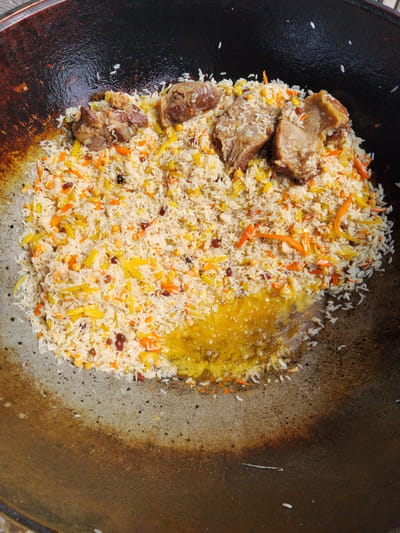 A top-down view shows a large, dark, round cooking vessel, possibly a kazan, filled with pilaf. The pilaf consists of white rice mixed with shredded carrots, chickpeas, raisins, and pieces of meat. Several chunks of browned meat are visible on top of the rice. A pool of golden oil is present in the lower right portion of the vessel, with some rice and vegetables submerged in it. The cooking vessel itself appears well-used, with a seasoned, dark surface. The scene is likely set outdoors, given the natural lighting. The image captures the preparation of a traditional Central Asian dish, potentially within the city of Ala-Buka, Kyrgyzstan, known for its cultural heritage and culinary traditions.
