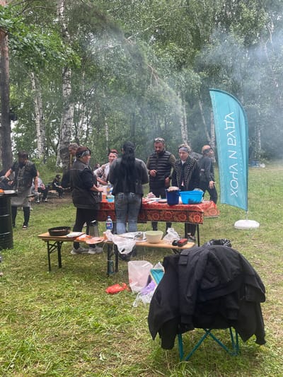 The image depicts an outdoor gathering in a wooded area, possibly a park or campground, on a cloudy day. Several individuals are present, engaged in what appears to be food preparation or a meal. A table covered with a patterned tablecloth is set up with various items, including bowls, food containers, and what looks like a grater. Some people are standing around the table, interacting with each other and the food. Smoke is visible in the background, suggesting a barbecue or campfire. A large teal flag with text is planted in the ground to the right of the scene. The ground is covered in grass and fallen leaves. The overall atmosphere suggests a casual, social event. The location is identified as Il'ichevskii, Russia.