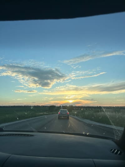 The image captures a view from inside a vehicle traveling on a road at sunset. The main subjects are the road, the sky, and a car in front. The scene is set in a rural landscape, with fields and trees lining the road. The sky is filled with wispy clouds and illuminated by the warm glow of the setting sun, indicating it is late afternoon or early evening. The weather appears clear, with the sun breaking through the clouds. The emotion conveyed is one of peaceful travel and the beauty of nature.

Notable details include the interior of the car, with a dashboard visible on the right side of the frame. The road ahead has white lines marking the lanes. The car in front has its taillights on, suggesting it is moving. The landscape on either side of the road is a mix of green fields and a distant treeline.

There is some visible text on the license plate of the car in front, which appears to be "AX68 60 AX".

The location context provided is Primortsi, Bulgaria, which helps to place the rural setting. The overall impression is a serene moment of a road trip during a beautiful sunset.