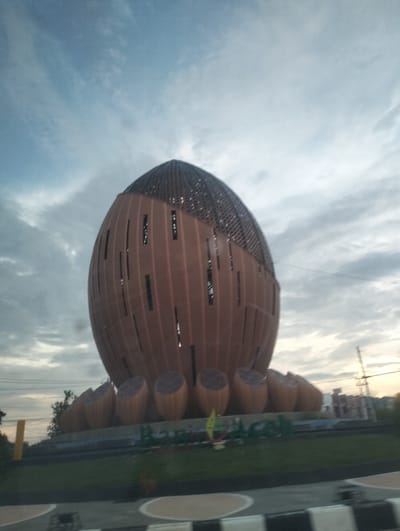 This media file depicts a large, spherical monument with a patterned, dark-colored dome-shaped roof. The main body of the monument is a reddish-brown color and features vertical, oblong windows. It is surrounded by several smaller, dome-shaped structures that appear to be part of the base. The monument is situated in a park-like setting with green grass and some trees visible in the background. In the far background, there are buildings and power lines, suggesting an urban or semi-urban environment.
The time of day appears to be either early morning or late evening due to the soft, diffused light and the gray, cloudy sky. The weather seems overcast, with the clouds obscuring the direct sun. The overall mood is calm and somewhat serene.
The inscription "RAMAH ACEH" is visible in large green letters on the ground in front of the monument. The monument's design, particularly its shape and the inscription, suggests it is a landmark or tourist attraction in the Aceh region of Indonesia. The context provided indicates the location is Sigli, Indonesia. There are no people visible in the frame, and the activity taking place is simply the monument standing as a static structure within its environment.