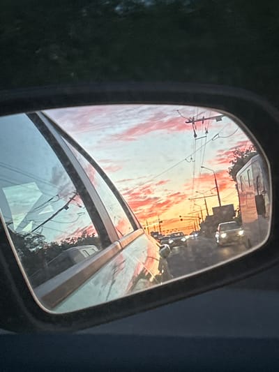 The image shows a car's side mirror reflecting a sunset view of Тимофеевка, Russia. Power lines and poles run along a street. Several vehicles are visible. The sunset creates a red and orange sky with clouds.