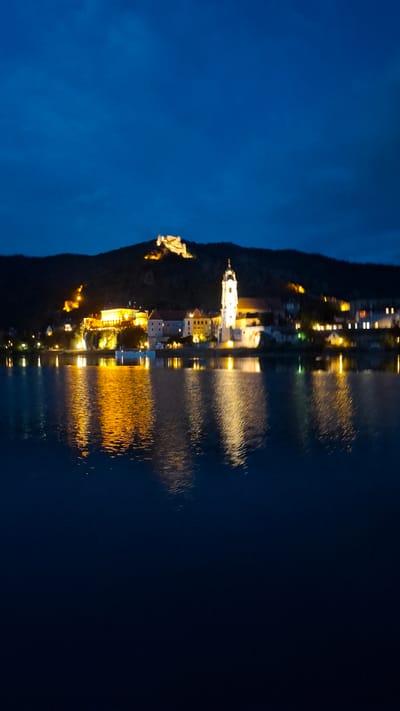 The image depicts the illuminated town of Rossatz-Arnsdorf, Austria, at dusk, as viewed across the Danube River. The sky is a deep blue, transitioning into a darker twilight. On the hillside overlooking the town, a large, illuminated castle ruin is visible. Below the castle, a cluster of buildings, including a prominent white church with a tall steeple, are brightly lit, casting warm yellow and white reflections onto the calm surface of the river. The reflections are elongated and somewhat distorted, typical of light on water. The overall scene conveys a serene and picturesque nighttime view of a historic Austrian town.