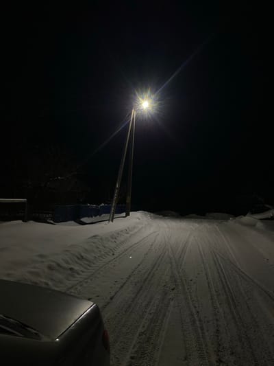 A nighttime scene features a bright street lamp illuminating a snow-covered road. Tire tracks are visible in the snow, leading away from the camera. The darkness dominates the left side of the frame. Part of a car is visible in the lower right corner. A fence is seen behind the snowbank. The light source creates a starburst effect.