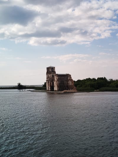 An abandoned, partially collapsed brick structure, resembling a church or bell tower, stands on a small island in a body of water. The building is weathered and in disrepair, with exposed brickwork and wooden scaffolding supporting parts of its structure. Lush green trees and reeds line the opposite bank and the island's edge. The sky above is partly cloudy with a mix of white and gray clouds. The water surface is rippled, reflecting the sky and the surrounding landscape. A few small, triangular structures, possibly buoys or markers, are visible in the water near the shore. The scene appears to be a historical site in Krokhino, Russia, known for its submerged or partially submerged architecture.