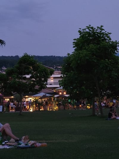 A nighttime view showcases a gathering spot with a building illuminated by warm lights. People are congregated both inside the structure and outside on the adjacent grass, some are seated on blankets. Trees provide a natural screen. A hill is visible in the distance. The sky is a dark, muted blue.