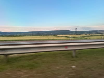 The image is a photograph of a rural landscape, possibly taken from a moving vehicle, with a highway guardrail in the foreground. The guardrail is made of metal and appears slightly worn. Beyond the guardrail, there is a grassy area, followed by a field of yellow crops. In the distance, there are rolling hills covered in green trees. The sky is a clear blue with some light clouds near the horizon. There are several tall power line towers visible in the distance. The scene appears to be set in a rural area, possibly the outskirts of Verkhnii Aktash, Russia, given the context provided. There are no people or specific actions depicted in the image. The overall impression is one of open space and natural scenery.