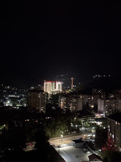 An aerial, night-time, vertical, medium shot captures a dimly lit cityscape in the Vardane Microdistrict, Russia. In the mid-ground, several multi-story buildings with illuminated windows are visible. One cluster of buildings in the distance is brightly lit from above and around its facades, with red lights outlining the rooftops. To the right of these buildings, a tall, red and white striped tower stands, illuminated with red lights at its peak. In the foreground, the dark silhouettes of trees and more buildings with illuminated windows are present. A road with a few cars and streetlights can be seen in the lower right portion of the frame. The sky is dark and unlit, with no visible celestial bodies. The overall scene depicts a residential area at night, with the exception of the prominent illuminated tower.