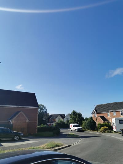 This is a wide-angle, eye-level, outdoor, daytime photograph of a residential street in Ripon, United Kingdom. The sky is a clear, bright blue with a faint, light-colored arc visible in the upper portion of the image. On the left side of the frame is the front of a two-story red-brick house with a dark-tiled roof. A dark-colored estate car is parked on the paved driveway in front of the house. A road curves through the middle of the image, with houses and trees lining the sides. On the right side of the road, there is a white van with a red logo on the side. Further down the road, another house with a dark-tiled roof is visible, and to its right, a white caravan with a bicycle rack is parked. The scene appears to be a quiet suburban neighborhood on a sunny day.