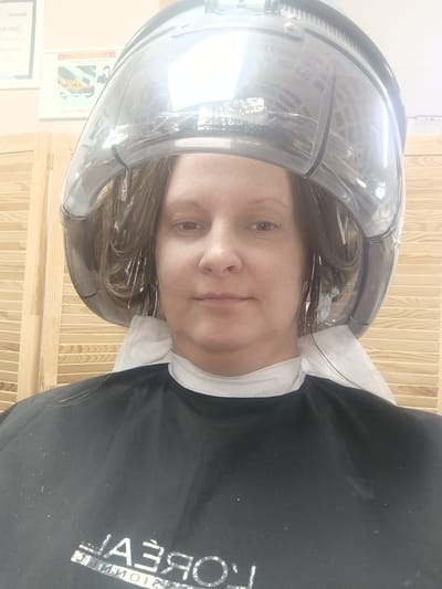 A woman is sitting under a hair dryer. She has foil in her hair, and is wearing a black salon cape with L'Oreal branding. The background includes a wooden screen and framed posters. The location is Closed administrative–territorial formation "Mezhgorye", Russia.