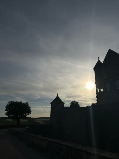 This forensic image captures the silhouette of a historical fortified structure, identified as located in Untergruppenbach, Germany, bathed in the light of a low sun during either sunrise or sunset. The sun, a bright, circular luminary, is positioned in the middle-right portion of the frame, generating a prominent vertical lens flare and a subtle green chromatic aberration in the upper-mid section.

The sky occupies the majority of the upper frame, displaying a gradient from darker grey-blue at the zenith to a lighter, warmer hue near the horizon. It features scattered, wispy clouds and multiple distinct airplane contrails.

Architectural elements dominate the mid-ground and right side. On the far right, a multi-storied stone building with a gabled roof and an attached smaller turret-like structure featuring a pointed roof is heavily shadowed, though some exterior wall sections and white-framed windows are visible on the sun-struck side. Towards the center-left, another conical-roofed stone tower stands, also silhouetted. A low, dark stone wall with a flat coping runs from the bottom center of the frame, extending towards the mid-left and bordering a dark, unidentifiable ground surface, possibly a road or path.

To the left of the central tower, a large, full-canopied deciduous tree is silhouetted against the lighter sky. In the distant background, rolling, cultivated fields and hills are discernible under the soft, ambient light. No human or animal subjects are visible, and no specific actions are taking place within the static scene.