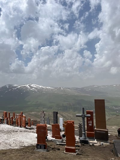 The image depicts a hillside cemetery in Hartagyugh, Armenia. Gravestones, predominantly made of brown stone, are arranged along the hillside. A large gray cross stands prominently among the graves. In the background, a green valley stretches towards a snow-capped mountain range. A cloudy sky occupies the upper left portion of the image. A single person is visible in the upper right corner of the scene.