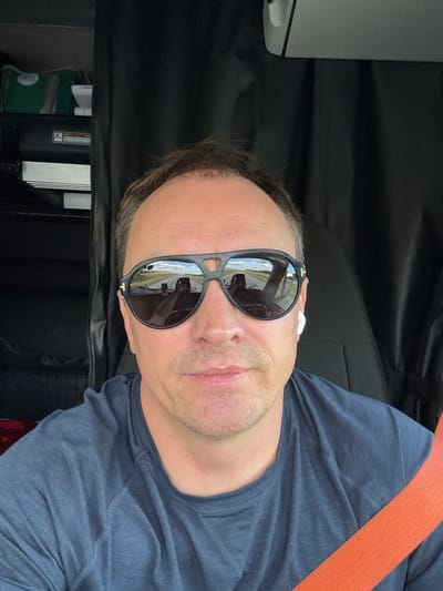 A man, wearing sunglasses and a navy blue t-shirt, is seated in the driver's seat of a vehicle. The sunglasses reflect a road with two other vehicles driving in the same direction, suggesting the man is in transit. A seatbelt, bright orange in color, is visible across his chest. He has a white wireless earbud in his right ear. In the background, there are dark curtains, and to the left, a shelf holds several items, including a green bag. The scene appears to be within the cabin of a truck. The location is Dola, United States.