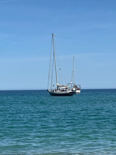 Two sailboats are moored in the clear blue waters of the Mediterranean Sea off the coast of Le Lavandou, France. The larger sailboat is in the foreground, positioned to the left of the frame, with its dark hull contrasting against the vibrant turquoise water. Its mast is tall and slender, with rigging clearly visible against the clear blue sky. The second sailboat is smaller and located slightly behind and to the right of the first, also a sailboat with a white hull and mast. Both vessels appear to be at anchor, stationary in the calm sea. The horizon line is sharp, dividing the deep blue of the sea from the lighter blue of the sky. A faint, distant landmass or coastline can be seen on the horizon. The foreground shows the gentle, rippling surface of the water.