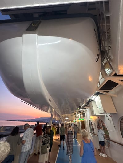 This media file captures a scene on the deck of a ship, likely a cruise ship, with a sunset visible in the background. The image is oriented sideways. In the foreground, a large white lifeboat, numbered "9", is prominently displayed. The text "NORWEGIAN NASSAU" is visible on the lifeboat, suggesting it belongs to a Norwegian cruise line.
A group of people are gathered on the deck, facing towards the water and the sunset. They appear to be tourists or passengers observing the view. Some individuals are casually dressed, while others are in more formal attire. The sunset casts a warm glow over the scene, with hues of pink, orange, and purple in the sky. The sea is visible in the distance, with faint lights suggesting a coastline or other vessels.
The overall mood appears relaxed and contemplative, as people take in the natural beauty of the sunset. The setting appears to be a port or a popular cruising route, given the presence of a lifeboat and the organized gathering of people. The weather seems pleasant, with clear skies contributing to the vibrant sunset. The time of day is clearly sunset. The location context of Motril, Spain, further solidifies the idea of a coastal cruise or stopover.