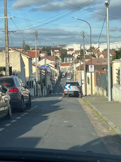 The image depicts a descending residential street in Argenteuil, France, observed from the interior of a vehicle. The foreground shows the top edge of a dark dashboard or windshield frame. On the left, portions of two parked dark-colored SUVs are visible; the closer one displays a silver roof rail and part of its rear fascia. White dashed lines mark the left side of the asphalt road, which narrows as it slopes downhill.

In the midground, a grey Renault Captur (license plate 990 BC) is parked on the right side of the street, facing away from the camera. Immediately behind it, a blue rectangular sign with a white left-pointing arrow is mounted on a pole. To the right of the road, a narrow, unpaved strip with sparse vegetation borders a concrete curb and a sidewalk. Numerous utility poles with overhead wires run along both sides of the street. A street lamp with a curved arm is visible on the right. Further down the hill on the right, a red circular "No Entry" sign is mounted on a pole, with an additional rectangular sign below it. Several detached or semi-detached residential houses with varied architectures, primarily featuring red-tiled pitched roofs and light-colored facades (cream, yellow, white), line both sides of the street. Additional vehicles, including a white van and other cars, are visible further down the road.

The background reveals a panorama of more residential buildings interspersed with trees. On the far right, a large, modern, multi-story white apartment complex dominates the distant skyline. A construction crane is visible on the left horizon. The sky is partially cloudy blue, indicating daylight conditions with direct sunlight casting distinct shadows across the road from the foreground vehicles. No people are visible in the scene.