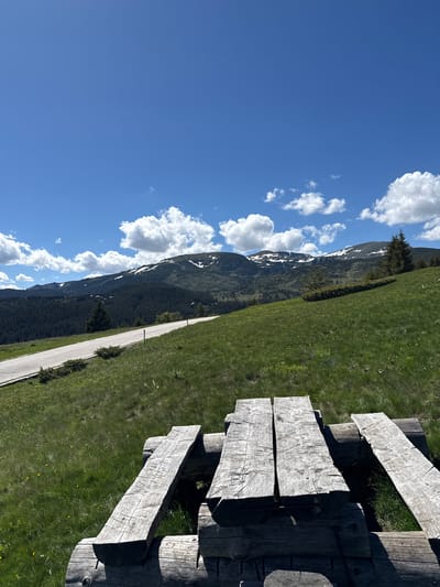 This is a photograph of a wooden picnic table in the foreground, with a paved road winding uphill through a grassy, mountainous landscape in the background. The sky is blue with scattered white clouds. The mountains in the distance are covered with dark green coniferous trees and patches of snow. The location is Cherna Mesta, Bulgaria, a mountain resort known for its natural beauty and outdoor activities. The image is taken from a slightly elevated perspective, looking towards the mountains. There are no people visible in the image.