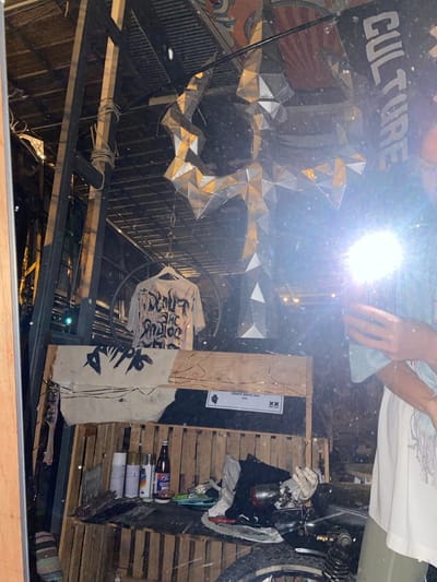 The image shows a cluttered, reflective interior space with visible ceiling beams and an irregular, metallic sculpture. A sign reading "CULTURE" is partially visible. A person holding a reflective object is present, creating a bright glare in the center. The space contains a motorcycle, spray paint cans, clothing, and wooden pallets. The location is Baan Nai Suan, Thailand.
